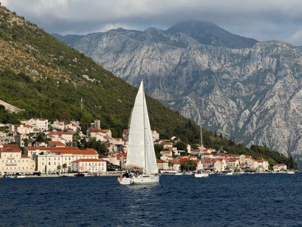 Sailboat gliding on the Adriatic Sea near Kotor, Montenegro, with terracotta-roofed coastal villages, ancient stone towers, and towering limestone cliffs rising dramatically in the background under a partly cloudy blue sky.